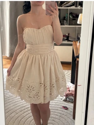 Beige axelbandslös kort klänning - Söt beige axelbandslös klänning med markerad midja och utställd kjol. Kjolen har fina broderade blommönster och vågig kant. Överdelen är veckad och ger en elegant look. Perfekt för sommarens alla tillfällen. Perfekt till midsommar💐💐obs jag på bild är xs/s så lite stor vid bysten😁