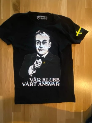 Svart AIK t-shirt med tryck - Svart t-shirt från Broder Tuck med stort tryck av en man i kostym och texten 'Vår klubb vårt ansvar' på framsidan. På ena ärmen finns ett gult kors, inspirerat av AIK:s färger. Perfekt för dig som vill visa klubbkänsla. från år 2014. Jätte sällsynt. Storlek S men passar tajt