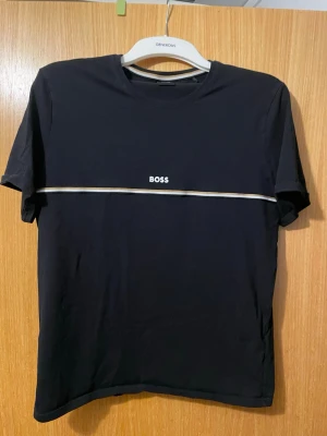 Svart BOSS t-shirt med vit rand - Snygg svart t-shirt från BOSS med vit och beige rand över bröstet och diskret BOSS-logga framtill. Klassisk rund halsringning och korta ärmar. Tillverkad i mjuk bomull för skön känsla och stilren look.
