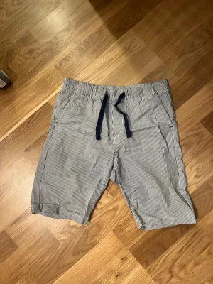 Randiga shorts med snörning barn. Strl 140 - Snygga randiga shorts med mörkblå snörning i midjan. De har smala mörkblå och vita ränder och är tillverkade i ett lätt och bekvämt bomullsmaterial. Perfekta för varma dagar och enkel att matcha med t-shirt eller linne.