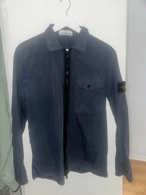 Mörkblå overshirt från Stone Island - Säljer en mörkblå overshirt från Stone Island med klassisk krage, dragkedja framtill och en bröstficka med knapp. Ikoniska Stone Island-märket på ärmen. Tillverkad i ett mjukt bomullsmaterial som ger en avslappnad men stilren look.