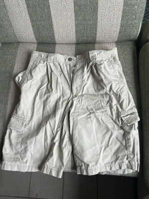 Beige cargoshorts med fickor - Säljer ett par beige cargoshorts med flera praktiska fickor på sidorna och baktill. Klassisk passform med knapp och dragkedja framtill. Perfekta för sommardagar och avslappnade outfits.