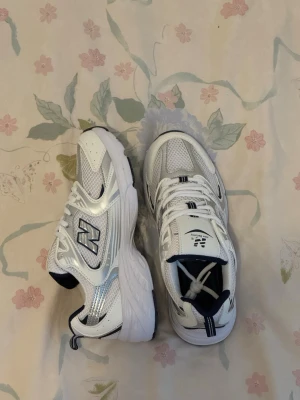 Vita New Balance 530 sneakers - Snygga vita New Balance 530 sneakers med silvriga och mörkblå detaljer. Skorna har en chunky sula och meshpaneler för extra ventilation. Klassisk logga på sidan och bekväm passform. Perfekta för dig som gillar retrostil och sportig vibe.