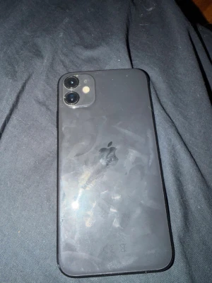 Svart iPhone 11 med dubbla kameror - Säljer en svart iPhone 11 med stilren design och rundade kanter. Baksidan är i glas och har två kameror samt Apple-loggan. Skärmen är stor och täcker nästan hela framsidan, perfekt för sociala medier och streaming.