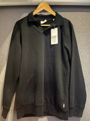Svart långärmad tröja Lindbergh - Stilren svart långärmad tröja från Lindbergh med klassisk krage och relaxed fit. Tröjan har ribbade muddar vid ärmslut och nederkant, och är tillverkad i ett mjukt material som passar perfekt till en avslappnad look.
