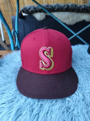 Vinröd keps med S-logga och jubileum - Snygg vinröd keps med stor S-logga i guld och vitt framtill. Skärmen är svart och på sidan finns ett broderat 20th Anniversary-märke med kompassmotiv. Klassisk fitted-modell i bomull, perfekt för dig som gillar sportig streetstyle.