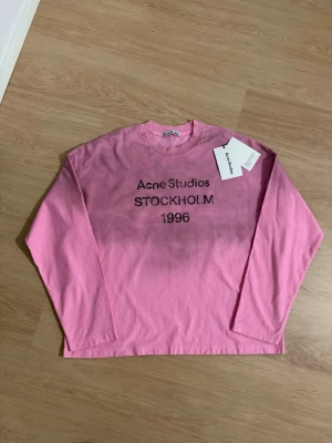 Rosa långärmad topp Acne Studios - Cool rosa långärmad topp från Acne Studios med tryck 'Acne Studios STOCKHOLM 1996' på bröstet. Klassisk rund halsringning och relaxed fit. Perfekt för dig som gillar streetwear och vill ha något unikt från ett ikoniskt svenskt märke.