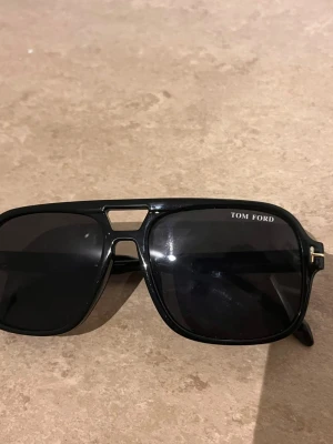 Svarta Tom Ford pilotbågar solglasögon - Snygga svarta solglasögon från Tom Ford med klassisk pilotform och bred båge. Glasen är mörka och bågarna har gulddetaljer vid gångjärnen. Perfekta för dig som vill ha en stilren och exklusiv look.