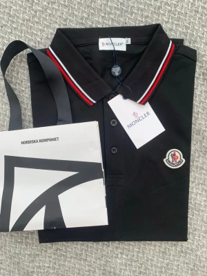 Svart Moncler pikéskjorta XL - Säljer en svart pikéskjorta från Moncler i storlek XL. Skjortan har klassisk krage med röda och vita ränder, knappar framtill och Moncler-logga på bröstet. Tillverkad i bomull för en skön känsla och stilren look.