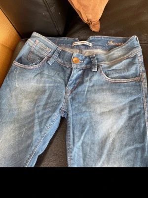Blå jeans från Fornania med slitningar - Säljer ett par blå jeans från Fornania med snygga slitningar och kontrastsömmar. Klassisk femficksmodell med orange knapp och detaljerade bakfickor. Jeansen har en rak passform och är tillverkade i denim med lätt tvättad look.