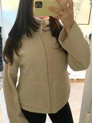 Beige teddyjacka  - Mysig beige teddyjacka med hög krage och dragkedja framtill. Jackan har två fickor och är gjord i fluffigt teddy-material som ger en avslappnad och trendig look. Perfekt för att hålla värmen under kyliga dagar.