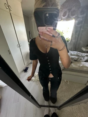 Svart topp med knappar och v-ringning - Snygg svart topp med v-ringning och dekorativa knappar längs hela framsidan. Toppen har korta ärmar och en liten slits nedtill framtill som ger en cool detalj. Passformen är slim och materialet känns mjukt och stretchigt.