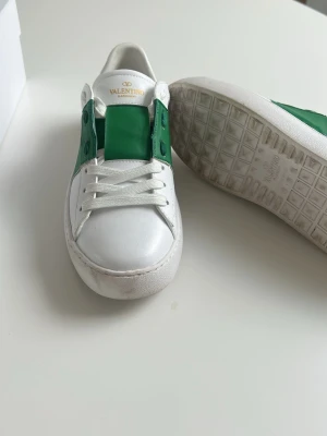 Valentino Garavani opens  - Snygga vita sneakers från Valentino Garavani med gröna detaljer på sidorna och ovandelen. Skorna har klassisk låg profil, vita skosnören och en robust gummisula. Tillverkade i läder för en exklusiv känsla och stilren look. Har använt bara tre gånger men dem var för små!!!