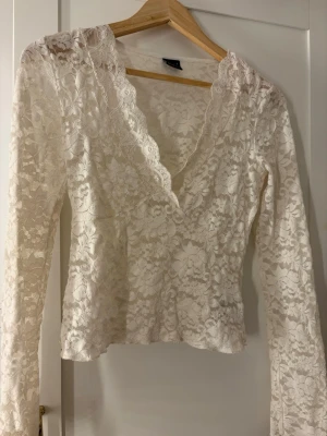 Vit spetsblus med v-ringning - Snygg vit blus i spets med v-ringad hals och långa ärmar. Blusen har ett romantiskt blommigt mönster och är perfekt för dig som gillar feminina detaljer. Passar till både jeans och kjol för en trendig look.
