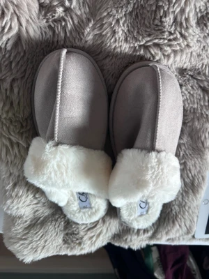 Gråa tofflor med vit fuskpäls - Mysiga gråa tofflor från Cecil med fluffig vit fuskpäls runt kanten och inuti. Tofflorna har en enkel slip-in design och är perfekta för att hålla fötterna varma hemma. Utsidan är i mjukt material som känns lyxigt och bekvämt.