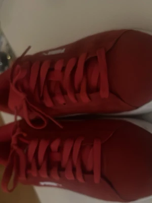 Röda Puma sneakers med vit sula - Snygga röda sneakers från Puma med klassisk vit sula och vit logga på sidan. Skorna har lågt skaft, röda skosnören och är tillverkade i syntetmaterial med detaljer i mockaimitation. Perfekta för dig som vill sticka ut med färg på fötterna.
