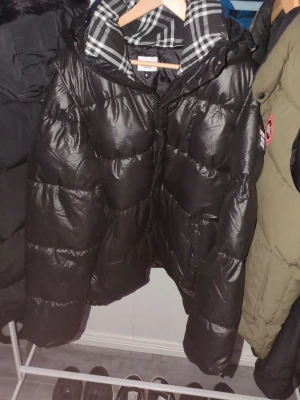 Svart dunjacka från Burberry - Säljer en svart dunjacka från Burberry med klassiskt rutigt foder i huvan och tryck på ärmen. Jackan har dragkedja, knappar och en stor huva. Perfekt för kalla vinterdagar och riktigt snygg streetstil. 