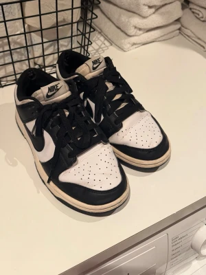 Nike panda Dunk Low svartvita sneakers - Svartvita Nike Dunk Low sneakers med klassisk design. Skorna har svart swoosh, svarta snören och vit tåbox med perforeringar. Yttermaterial i läder och låg siluett som ger en clean och tidlös look. Perfekta för dig som gillar streetwear och sneakerskultur.