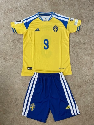 Sverige fotbollströja och shorts Adidas - Säljer ett matchställ från Sveriges landslag med gul tröja och blå shorts. Tröjan har nummer 9 och ISAK på ryggen, samt landslagets emblem och Adidas-logga. Shortsen är blå med vita ränder och har också landslagets emblem. Materialet är lätt och andas, perfekt för fotboll.