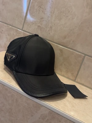 Svart keps från Prada i nylon - Snygg svart keps från Prada i glansigt nylonmaterial med klassisk böjd skärm och ikonisk metallogo på sidan. Kepsen har en stilren design och justerbar passform. Perfekt accessoar för en trendig och sportig look.