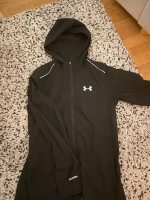 Svart hoodie från Under Armour - Snygg svart hoodie från Under Armour med dragkedja framtill och vit logga på bröstet. Stora fickor på sidorna och huva. På ryggen finns en stor vit UNDER ARMOUR-text. Tillverkad i ett mjukt och stretchigt material, perfekt för träning eller chill.