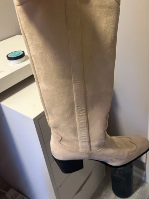 Beige höga cowboy boots i mocka - Snygga beige cowboy boots i mocka med spetsig tå och bred klack. Lite sliten under till men annars syns det inte att den är använd 