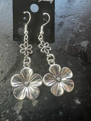 Silvriga blommiga hängörhängen - Säljer ett par silvriga hängörhängen i metall med två blommor på varje örhänge. Den övre blomman är mindre och den undre större, båda med fina detaljerade kronblad. Perfekta för dig som gillar statement-smycken med blommig design.