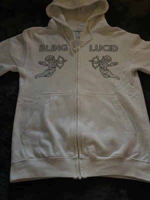 Vit hoodie från Bling Lucid - Vit zip-hoodie från BLING LUCID med huva och fickor framtill. På bröstet finns två änglamotiv och texten 'BLING LUCID' i glittriga stenar. Tillverkad i mjukt material, perfekt för en chill och trendig look.