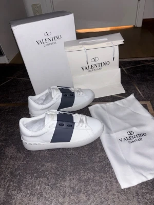 Valentino Garavani Open sneakers - Säljer ett par Valentino Garavani Open sneakers i vitt kalvskinn med en blå detalj. Klassisk och stilren design med rund tå och platt sula. Perfekta för dig som vill ha exklusiva och trendiga sneakers från Valentino. Storlek 43