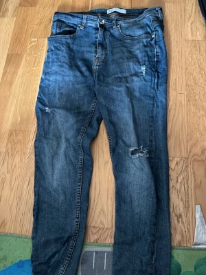 Blå slitna jeans med hål - Säljer ett par blå jeans med slitningar och hål på låret och knät. Jeansen har klassisk femficksdesign, knappgylf och en avslappnad passform. Perfekta för en casual och trendig look.