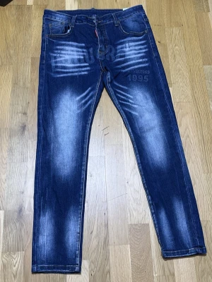  jeans - Säljer ett par blå jeans med coola slitningar och tryckdetaljer både fram och bak. Jeansen har raka ben, klassisk femficksmodell och tydliga vita tvätteffekter. Materialet är denim i bomull och de har trycket 'Established 1995' på framsidan.