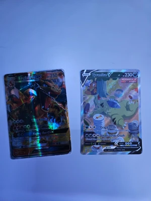 Pokémonkort Charizard GX & Tyranitar V - Två holografiska Pokémonkort: Charizard GX med färgstark design och Tyranitar V med motiv av Tyranitar som vilar bland matrester. Båda korten har metallic-effekt och detaljerade illustrationer. Perfekt för samlare eller Pokémonfans.
