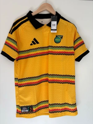 Jamaica matchtröja Adidas gul - Säljer en gul Jamaica matchtröja från Adidas med svarta, gröna och röda detaljer. Tröjan har svart krage, svarta muddar och tre svarta ränder på axlarna. Broderad Jamaica-logga på bröstet och texten 'Reggae Boyz' på ryggen. Tillverkad i lätt och ventilerande material.