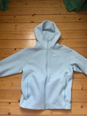 Ljusblå Nike Nocta hoodie med dragkedja - Snygg ljusblå hoodie från Nike med hel dragkedja framtill och två fickor med dragkedja. Klassisk Nike-logga på bröstet och huva för extra stilpoäng. Perfekt för dig som gillar sportig och clean look.