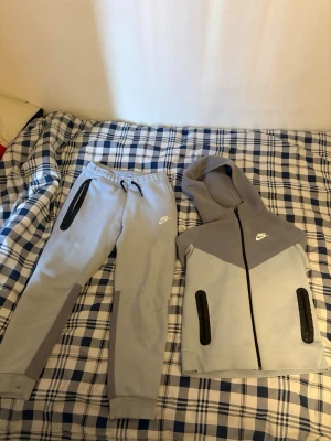 Nike Tech Fleece mjukisbyxor och hoodie  - Säljer ett par Nike Tech Fleece mjukisbyxor i ljusgrått med mörkgrå detaljer och svarta dragkedjor. Byxorna har en slim fit, resår i midjan med snörning och fickor med dragkedja. hoodie har samma färg som byxor och har dragdjedja på båda sidorna. Perfekta för chill eller träning.  Hoddien är 147-158 cm och samma ska med byxor