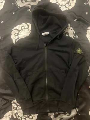 Stone Island Zip Hoodie Svart - Säljer en klassisk svart Stone Island zip hoodie. Tröjan är sparsamt använd och i bra skick.  Färg: Svart.  Storlek: M. (För referens är jag 181cm och den sitter fint)  Autenticitet: 100% äkta. Certilogo/QR-kod finns och kan skickas i DM till seriösa köpare för verifiering. Kvitto finns ej kvar.  Märke: Patchen på ärmen är i fint skick. Finns för upphämtning i Gnesta eller skickas via posten samma dag som du lägger beställningen. Kan gå ner i pris vid snabb och smidig affär! Skicka DM vid frågor