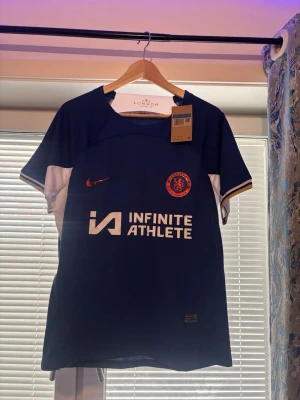 Chelsea FC matchtröja blå Nike - Chelsea FC matchtröja i blått med vita detaljer och guldkant på ärmarna. Nike-logga och Chelsea-märke på bröstet samt Infinite Athlete-tryck på magen. Tillverkad i lätt och ventilerande material, perfekt för träning eller match.