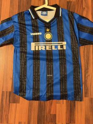 Inter Milan retro tröja 90-talet - Klassisk Inter Milan matchtröja från Umbro med blå och svarta vertikala ränder, Pirelli-logga på bröstet och klubbmärke med stjärna. Tröjan har krage med vit och svart detalj och är tillverkad i lätt, ventilerande material. Perfekt för fotbollsfans!