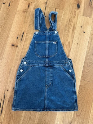 Blå denim jumpsuit från Tommy Jeans - Cool blå denim jumpsuit från Tommy Jeans med klassiska hängslen och stora fickor fram. Snygga detaljer som logotyp på bröstfickan och insidan. Perfekt för dig som gillar streetstyle och vill ha något bekvämt och trendigt.