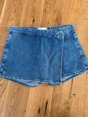 Blå denim skort med omlott - Säljer en blå denim skort med omlott-design och klassisk jeanslook. Skorten har en rak passform och är tillverkad i slitstarkt bomullsdenim. Perfekt för dig som vill ha en cool och avslappnad stil.