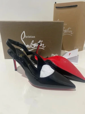 Svarta lackpumps från Louboutin - Snygga svarta pumps i lack från Christian Louboutin med klassisk röd sula och spetsig tå. Skorna har öppen häl med rem och smal klack som ger en elegant siluett. Perfekta för dig som vill ha en statement-sko med lyxig känsla.