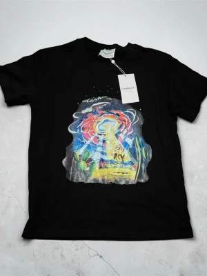 Svart t-shirt med färgglatt tryck Casablanca - Svart t-shirt från Casablanca med ett stort, färgglatt tryck framtill som visar en surrealistisk scen med kaktusar, UFO och starka färger. Klassisk rund hals och korta ärmar. Perfekt för dig som vill sticka ut med en unik design.