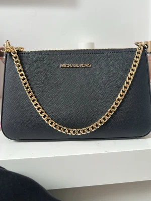 Svart Michael Kors handväska med kedja - Stilren svart handväska från Michael Kors i strukturerat läder med guldfärgad kedjerem och dragkedja. Väskan har ett diskret logotypmärke framtill och ett fodrat innerfack med ficka. Perfekt för dig som gillar klassisk och lyxig design.Man får även med ett annat alextrem i svart som är lite längre så man kan ha väskan lite längre ner om det är önskat. 
