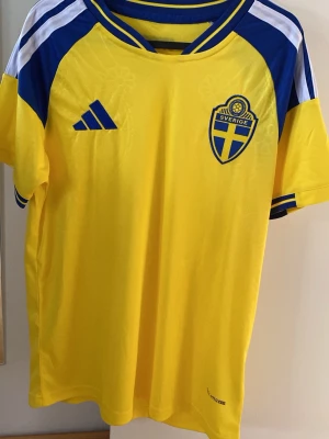 Svenska landslagströja Adidas gul - Gul svensk landslagströja från Afidas. Tröjan har Sveriges landslagsemblem på bröstet och Adidas-logga. Tillverkad i lätt och ventilerande funktionsmaterial, perfekt för fotboll eller träning.