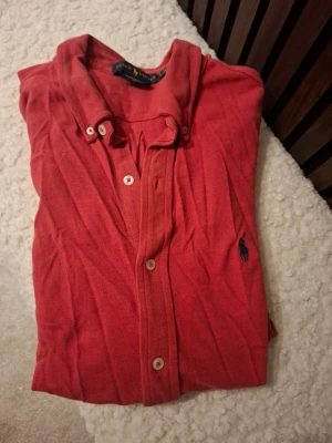 Röd skjorta från Ralph Lauren - Klassisk röd skjorta från Ralph Lauren med knappar framtill och ikoniska loggan broderad på bröstet. Skjortan har krage och är tillverkad i bomull för en bekväm känsla. Perfekt för dig som gillar stilrena plagg med en sportig touch.