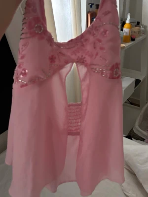 Rosa babydoll med broderi och paljetter - Söt rosa babydoll i transparent mesh med broderade blommor och paljettdetaljer upptill. Halterneck-modell med öppen rygg och elastisk detalj bak. Lätt och luftig känsla, perfekt för att känna sig extra fin.