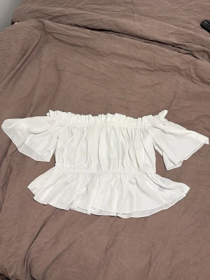 Vit offshoulder volangblus - Söt vit offshoulder blus med volangkant och korta vida ärmar. Toppen har resår vid axlar och midja för en bekväm passform och en flowy siluett. Perfekt för en somrig och trendig look.