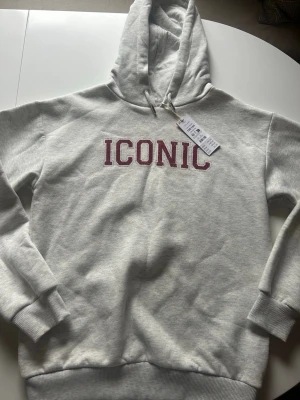 Ljusgrå hoodie med röd text Gina tricot - Säljer en ljusgrå hoodie från Gina tricot med röd texten 'ICONIC' på bröstet. Hoodien har en klassisk huva och är gjord i mjukt bomullsmaterial. Aldrig använd, lappen sitter kvar.