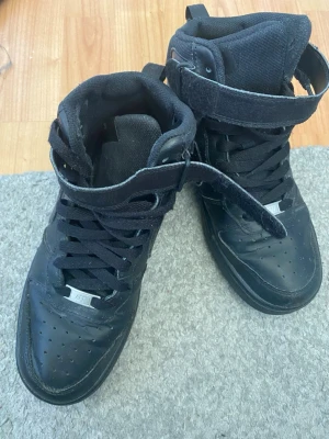Svarta Nike Air Force  High sneakers - Säljer ett par klassiska svarta Nike Air Force 1 High sneakers med högt skaft, snörning och kardborreband runt ankeln. Skorna är i helsvart läder med diskret Nike-logga på sidan och perforerade detaljer på tån. Perfekta för en streetwear-look.