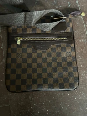 Brun H&M armväska - Snygg brun ryggsäck från H&M med klassiskt rutigt Damier-mönster och ikoniskt mönstrat foder. Väskan har dragkedja i metall och justerbara axelband i textil. Perfekt för dig som vill ha en stilren och lyxig accessoar till vardagen. Det enda med den är att den är lite sönder i kedjan men alla delar ingår så den går att fixa